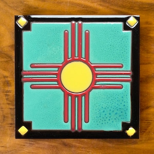 May include: Square ceramic tile featuring a turquoise background with a central yellow sun symbol and red rays. The tile is framed by a black border with yellow diamond accents in each corner. The design is a Southwestern motif.