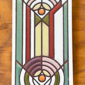 May include: A white ceramic tile with a geometric design in shades of red, green, yellow, pink, and purple. The design features concentric circles and triangles.