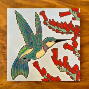 May include: A ceramic tile with a hand-painted design of a green and blue hummingbird with a long beak, hovering near red flowers. The tile has a white background.