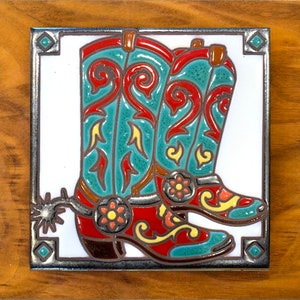 May include: A ceramic tile with a white background and a silver border features a pair of red and turquoise cowboy boots with intricate floral designs. The boots have a spur on the heel of one boot.
