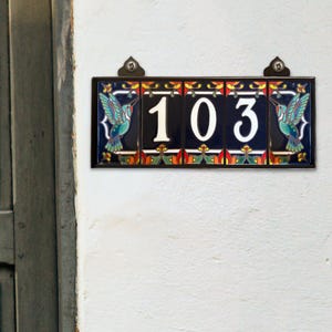 May include: A ceramic house number sign with the number 103 in white on a blue background. The sign is decorated with colorful floral designs and two hummingbirds on either side of the number.
