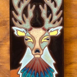 May include: A ceramic tile with a stylized image of a deer with large antlers. The deer is painted in shades of brown, yellow, blue, and green on a black background.