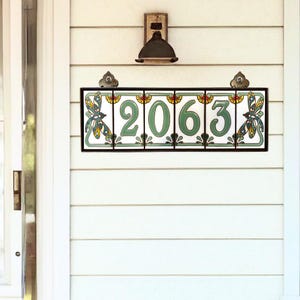 May include: A white ceramic house number sign with black framing and green and blue floral accents. The numbers 2063 are displayed in black on a white background.