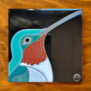 May include: A ceramic tile with a colorful illustration of a hummingbird with a long beak, red throat, and teal blue body. The tile has a black background and a small logo in the bottom right corner.