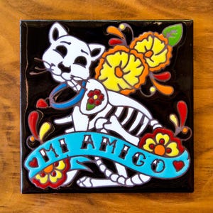 May include: Square ceramic tile with a black background featuring a white cat skeleton design. The cat holds a blue ribbon with the words "MI AMIGO" in turquoise. The design includes yellow, orange, and red floral accents.