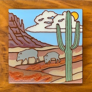 Puede incluir: Un azulejo de cerámica con una escena de desierto que presenta dos javelinas grises caminando en la arena, un alto cactus saguaro verde, un cielo azul con nubes blancas y una montaña marrón en el fondo.