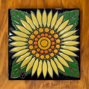 May include: A ceramic tile with a yellow sunflower design. The sunflower has a yellow center with orange and brown dots. The sunflower is surrounded by green leaves.