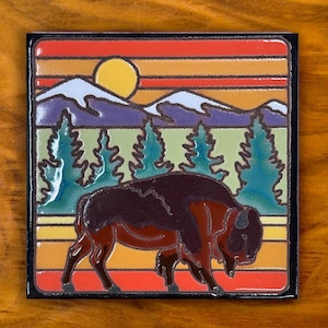 May include: Square tile featuring a bison walking across a landscape. The design includes a sunset with a yellow sun, mountains, and evergreen trees. The bison is brown and red, set against a colorful background.