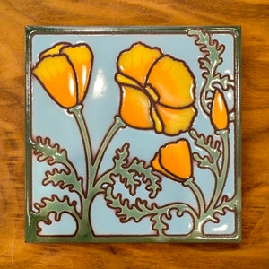 May include: A blue ceramic tile with a stylized design of orange California poppies with green leaves. The tile has a glossy finish.