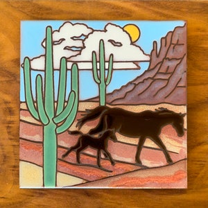 May include: A ceramic tile with a desert scene featuring a brown and black horse and foal running through a desert landscape with green cacti and a blue sky with white clouds.