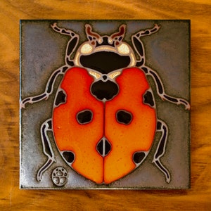May include: A ceramic tile with a hand-painted image of a ladybug. The ladybug is red with black spots and has a black head with white markings. The tile has a metallic gray background.