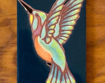 May include: A ceramic tile with a blue background and a colorful hummingbird illustration. The hummingbird is flying with its wings spread and has a long beak. The tile is a decorative piece for a home or garden.
