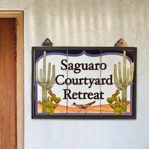May include: Rectangular tile sign with the words "Saguaro Courtyard Retreat" in black lettering. The sign features illustrations of cacti and a roadrunner on a desert landscape background. The sign is framed in a dark brown border.