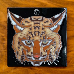 May include: A black and white ceramic tile with a detailed illustration of a bobcat's face. The bobcat's fur is depicted in shades of brown, orange, and white. The tile has a black background and a white circle with a symbol in the center.