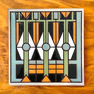 May include: A square ceramic tile with a geometric design in shades of green, orange, black, and white. The design features a repeating pattern of diamonds and triangles.