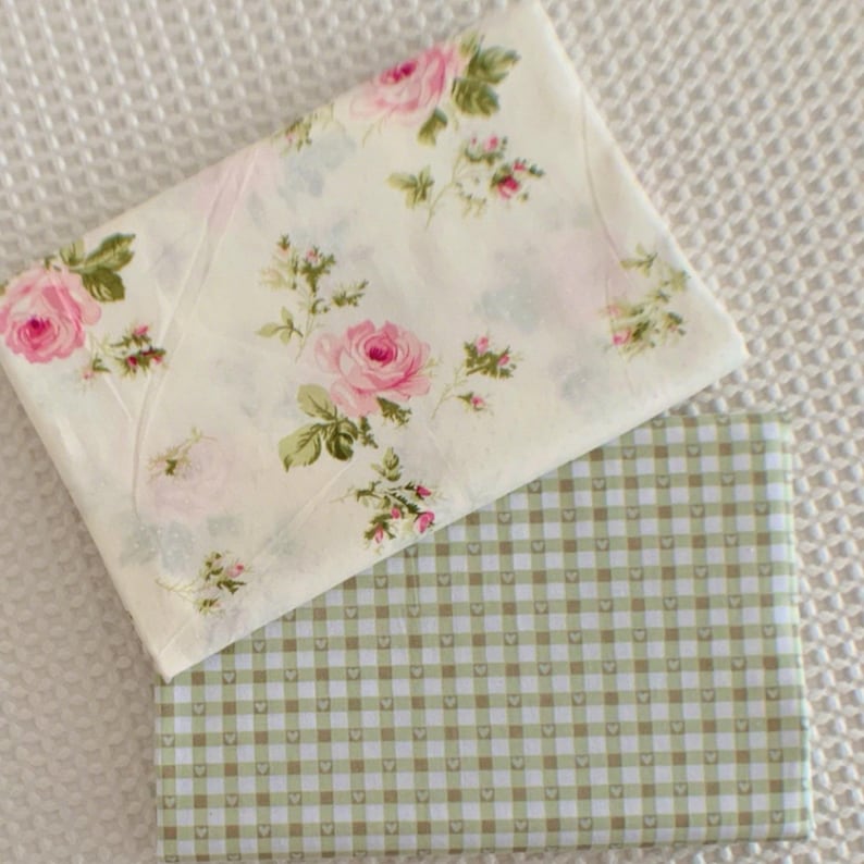May include: Two folded fabric squares. The top square features a cream background with pink roses and green leaves. The bottom square has a green and white checkered pattern with small heart shapes in the squares.