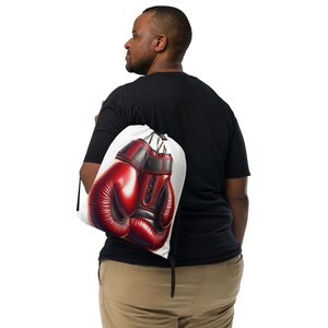 May include: A white drawstring bag featuring a vibrant illustration of red and black boxing gloves. The bag is carried over the shoulder, showcasing the detailed design of the gloves, which have a glossy finish.