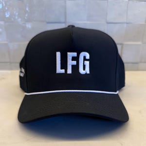 May include: Black trucker hat featuring the embroidered letters "LFG" in white. The hat has a white rope detail above the brim and a curved peak. The hat is made of a durable material and is suitable for outdoor activities.