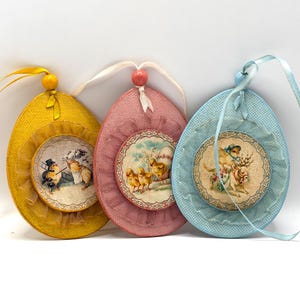 Pode incluir: Três enfeites de ovo de Páscoa em amarelo, rosa e azul. Cada ovo apresenta uma imagem circular em estilo vintage com uma borda decorativa e uma fita para pendurar. Os enfeites têm uma guarnição com babados e uma conta no topo.