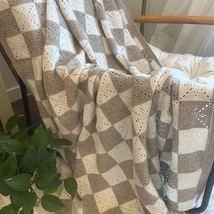 Puede incluir: Una manta de ganchillo hecha a mano en una paleta de colores neutros crema y topo. La manta presenta un patrón de cuadros con motivos cuadrados y un borde decorativo. La manta está sobre una silla, mostrando su textura y diseño.