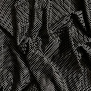 May include: A black fabric featuring a repeating, raised, small-scale grid or honeycomb texture. The material is draped, creating deep folds and shadows that highlight the consistent geometric pattern across the entire surface of the textile.
