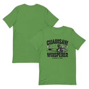 May include: A grass green short-sleeve t-shirt featuring a black graphic of a chainsaw centred on the chest. The text reads "CHAINSAW WHISPERER" in bold letters above and below the chainsaw, with "USA CERTIFIED - EST. 2008" printed below.