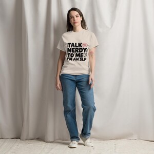 以下が含まれることがあります： ベージュの半袖Tシャツ。黒い文字で「TALK NERDY TO ME I'M AN SLP」と書かれ、赤い音波アイコンと吹き出しのグラフィックが添えられています。青いデニムジーンズと白いスニーカーを合わせています。