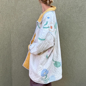 May include: Cream-colored quilted coat with floral and leaf appliqués in blue, green, and orange. The coat has a yellow collar and lining. The coat is knee-length and appears to be handmade.