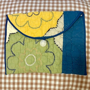 May include: A quilted clutch with a floral design. The clutch features a combination of yellow, green, and blue fabric, with a white button closure. The floral pattern is created with embroidered details.