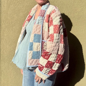 May include: A quilted jacket featuring a patchwork design of red, white, and blue fabric squares with various small-scale patterns. The garment has long sleeves, a light blue trim along the front opening and cuffs, and a boxy, open-front silhouette.