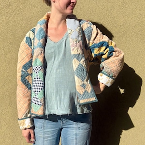 May include: A long-sleeve jacket constructed from quilted fabric featuring a patchwork design of various patterns including plaid, gingham, and floral prints in shades of blue, yellow, peach, and white, worn over a light blue v-neck top and jeans.