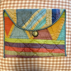 May include: A colorful, quilted clutch with a geometric design. The clutch features a variety of colors, including orange, blue, yellow, green, and purple. It has a rounded flap secured with a brass-colored button and is trimmed with a teal-colored piping.