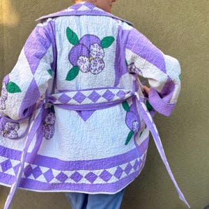 May include: A quilted jacket featuring a white base with lavender floral pansy appliqués and green leaves. The garment includes a matching tie belt, a diamond-patterned border along the hem, and long sleeves with folded cuffs, showcasing intricate stitching throughout.