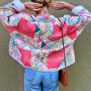 May include: A quilted jacket in a patchwork design, featuring a mix of pink, blue, green, and yellow fabrics. The jacket has a rounded collar and cuffs with white fabric. The jacket is worn with blue jeans and a brown shoulder bag.