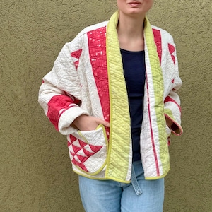 May include: A quilted jacket in a patchwork design. The jacket features a cream-colored base with red and white geometric patterns. The edges are trimmed with a bright yellow-green fabric. The jacket has pockets and a relaxed fit.