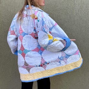 May include: A quilted jacket featuring a patchwork design with large white circles and colorful fabric squares. The jacket has a blue collar and cuffs, with a yellow and blue border along the hem. The overall style is vintage and handmade.