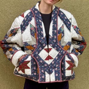 May include: A quilted jacket with a patchwork design. The jacket features a white base with colorful floral and geometric patterns in blue, red, and orange. The edges are trimmed with a pink fabric. It has pockets and a collar.