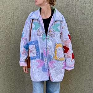 May include: A white quilted jacket with a patchwork design featuring various colors and patterns. The jacket has a collar, pockets, and a button closure. The quilt design includes pink, blue, red, and yellow patches. The jacket is worn over a black top and blue jeans.