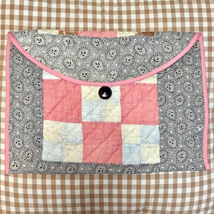 May include: A quilted fabric envelope with a pink trim and a black button closure. The envelope features a patchwork design with pink, white, and blue squares, set against a black and white floral pattern.