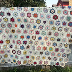 May include: A handmade quilt featuring a repeating pattern of multi-colored hexagonal fabric patches arranged on a cream-colored background. The quilt has a scalloped edge and displays a variety of floral and geometric prints in red, blue, yellow, pink, and green hues.
