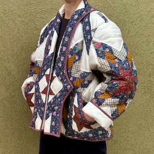 May include: A quilted jacket featuring a patchwork design with white panels and various floral and geometric patterns in shades of blue, red, and gold. The garment has long sleeves, a shawl collar, and a front opening, constructed with visible stitching.