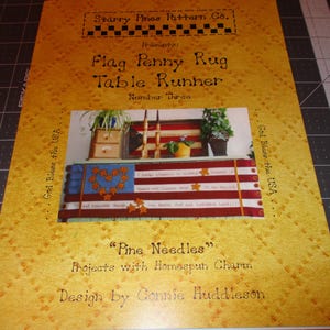 May include: A yellow cover with a title that reads "Flag Penny Rug Table Runner" by Starry Pines Pattern Co. The cover features a photo of a table runner with a patriotic theme. The text "God Bless the USA" is on the left and right sides of the cover. The text "Pine Needles" is below the photo. The text "Projects with Homespun Charm" is below "Pine Needles". The text "Design by Connie Huddleson" is at the bottom of the cover.