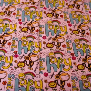May include: Pink fabric with a repeating pattern of smiling monkeys, suns, rainbows, and the word "Happy" in blue and yellow letters. The fabric has a white background and is decorated with small pink flowers.