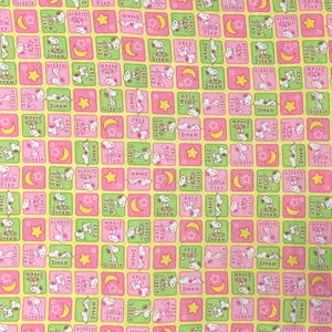 May include: A repeating pattern fabric featuring Snoopy and friends in pink and green squares on a yellow background. The squares contain Snoopy, stars, moons, and the words "Dream", "Play", and "Giggle".