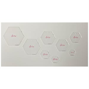 May include: A collection of clear acrylic hexagon quilting templates of varying sizes. Each template has a small hole and a red logo. The templates are arranged on a light gray background.