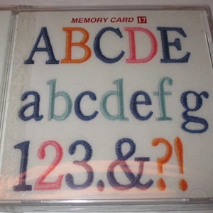 May include: A white memory card case with the text "MEMORY CARD 17" printed on the front. The case shows a colourful embroidered alphabet, including uppercase letters A through E, lowercase letters a through g, and numbers 1 through 3, along with the symbols & and ?!