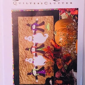 May include: A Halloween-themed quilt pattern featuring a row of witches and ghosts holding hands. The quilt is surrounded by fall foliage and a large orange pumpkin. The text "Halloween Ho-Down QC235-10" is printed below the quilt.