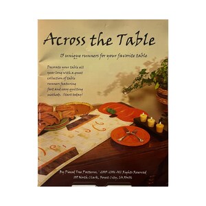 May include: A book cover titled "Across the Table" with text describing 15 unique table runners. The image shows a table setting with orange plates, candles, and a runner with decorative elements.