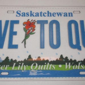 May include: A blue and white Saskatchewan license plate with the text "I Live To Quilt" and a red tiger lily. The bottom of the plate reads "Tiger Lily Quilts - Wolseley".