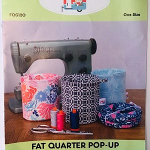 May include: A sewing pattern for a pop-up container, featuring a vintage sewing machine, spools of thread, scissors, and three fabric containers with different patterns. The pattern is titled "Fat Quarter Pop-Up" and includes instructions, a spring, a label, and elastic. The pattern is from "The Fat Quarter Gypsy".